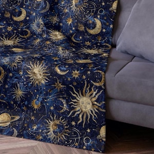 May include: A navy blue throw blanket with a celestial design featuring gold suns, moons, stars, and planets. The blanket is draped over a gray sofa, showcasing the intricate pattern and soft texture.