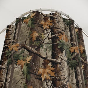May include: A camouflage shower curtain featuring a forest scene with brown tree trunks, branches, and green and orange leaves. The curtain hangs from a curved, silver-colored shower rod.