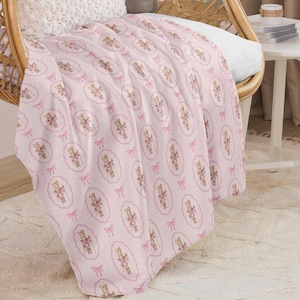 May include: A pink blanket featuring a repeating pattern of toy soldiers within oval frames, interspersed with pink bows. The blanket is draped over a wicker chair, offering a cosy and decorative appearance. The blanket is made of a soft material.