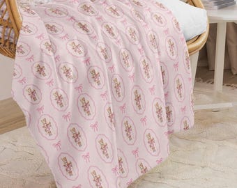 May include: A pink blanket with a repeating pattern of a toy soldier in an oval frame, accented with pink bows. The blanket is draped over a wicker chair, creating a cozy and decorative look. The blanket is made of a soft material.
