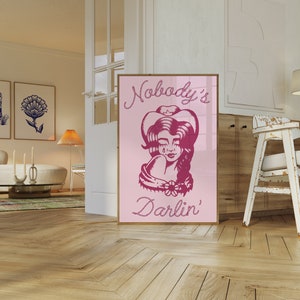 May include: Pink and white poster featuring a drawing of a woman wearing a cowboy hat and a bandana. The text on the poster reads "Nobody's Darlin'" in a rope-like font.