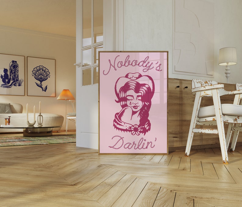 May include: Pink and white poster featuring a drawing of a woman wearing a cowboy hat and a bandana. The text on the poster reads "Nobody's Darlin'" in a rope-like font.