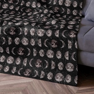 Può includere: Una coperta nera con un motivo ripetuto delle fasi lunari in tonalità di grigio e bianco. La coperta è drappeggiata su un divano grigio, mostrando il design celeste. Il motivo include lune piene, crescenti e altre fasi lunari.