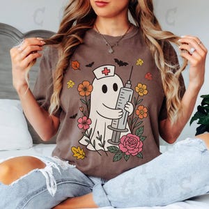 May include: Brown t-shirt featuring a cartoon ghost wearing a nurse's hat and holding a syringe. The ghost is surrounded by flowers, bats, and autumn leaves. The design is printed on a brown t-shirt.