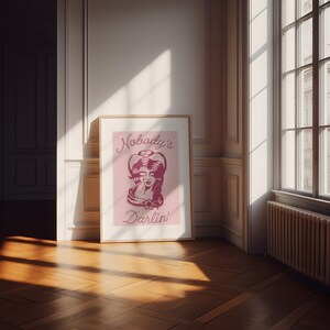 May include: A framed pink poster with a drawing of a woman wearing a cowboy hat. The text on the poster reads "Nobody's Darlin'".