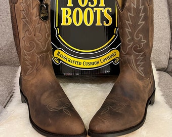May include: A pair of brown leather cowboy boots with intricate stitching and a pointed toe. The boots are sitting on a white fluffy surface and are next to a box that says "Dan Post Boots" with the slogan "Handcrafted Cushion Comfort".