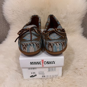 Könnte beinhalten: Ein Paar Minnetonka Baja Moc Turquoise Mokassins. Die Schuhe haben ein gewebtes Muster in Blau, Braun und Weiß mit Lederschnüren. Die Schuhe sind Größe 41 und stehen auf dem Schuhkarton.