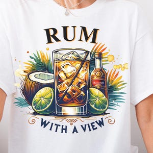 Op de afbeelding: Wit T-shirt met een afbeelding van een tropisch drankje met een limoenpartje, een kokosnoot en palmbladeren. De tekst "RUM WITH A VIEW" staat onder het drankje.