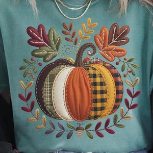 May include: Teal-colored t-shirt featuring a patchwork pumpkin design. The pumpkin has sections of orange, yellow plaid, and white fabric. Surrounding the pumpkin are embroidered leaves and berries, creating a fall-themed graphic.