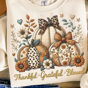 May include: Cream-colored sweatshirt featuring a fall-themed pumpkin design with floral accents and the words "Thankful Grateful Blessed." The pumpkins have various patterns and bows. The sweatshirt is paired with blue jeans and a brown hat.