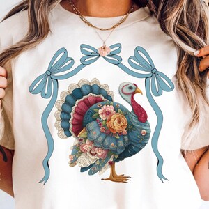 May include: Cream-colored t-shirt featuring a detailed illustration of a turkey adorned with flowers and lace. The turkey has a blue and red color scheme, with a floral design. Two light blue bows are at the top. The shirt is a Thanksgiving-themed design.