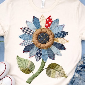May include: Cream-colored t-shirt featuring a patchwork sunflower design. The sunflower has denim, red star, and patterned fabric petals, a brown center, and green leaves and stem. The shirt is paired with jeans and white sneakers.
