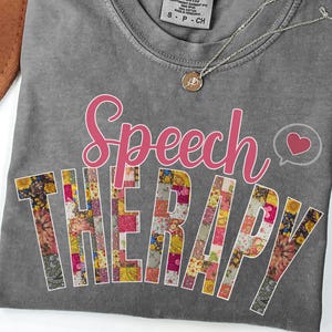 May include: A gray Comfort Colors t-shirt with the words "Speech Therapy" in pink script. The word "THERAPY" is filled with a floral patchwork design. A silver necklace with a pendant is draped on the shirt.