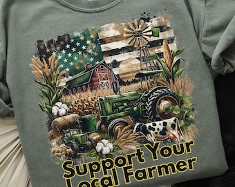 Könnte beinhalten: Salbeigrünes T-Shirt mit einer Grafik, die einen grünen Traktor, die amerikanische Flagge, eine Scheune, eine Windmühle und den Text "Support Your Local Farmer" zeigt. Das Design umfasst Mais, Baumwolle und eine Kuh.
