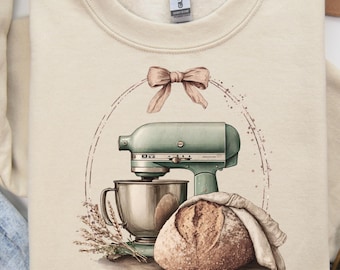 May include: Cream-colored sweatshirt featuring a watercolor illustration of a vintage mint green stand mixer, a loaf of bread, and a bow. The design includes a silver mixing bowl, a sprig of lavender, and a bread covered with a cloth.