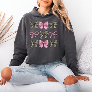 May include: A charcoal gray hooded sweatshirt featuring a graphic print of nine bows arranged in a grid. The bows alternate between solid light pink and a camouflage pattern in shades of green, brown, and black. The hoodie has long sleeves and a hood.