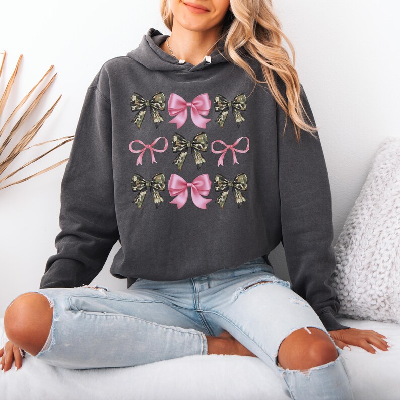 May include: A charcoal gray hooded sweatshirt featuring a graphic print of nine bows arranged in a grid. The bows alternate between solid light pink and a camouflage pattern in shades of green, brown, and black. The hoodie has long sleeves and a hood.