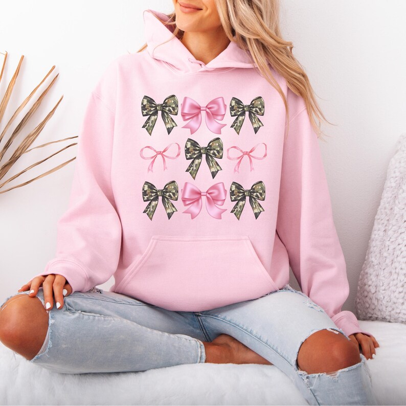 May include: A light pink hooded sweatshirt featuring a repeating graphic pattern of bows. The design includes solid pink bows and camouflage-patterned bows arranged in a grid across the front, with a large kangaroo pocket at the waist.
