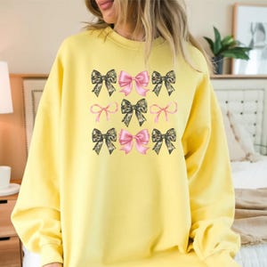 May include: A bright yellow long-sleeve sweatshirt featuring a central graphic print of nine bows arranged in a three-by-three grid. The bows alternate between solid pink and a camouflage pattern in shades of green, brown, and black.
