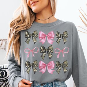 May include: A grey long-sleeve sweatshirt featuring a repeating grid pattern of graphic bows. The design includes alternating light pink bows and camouflage-patterned bows arranged in three rows across the front of the garment.