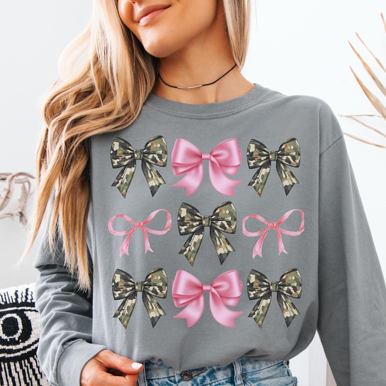May include: A grey long-sleeve sweatshirt featuring a repeating grid pattern of graphic bows. The design includes alternating light pink bows and camouflage-patterned bows arranged in three rows across the front of the garment.