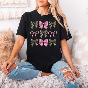 May include: A black short-sleeved t-shirt featuring a graphic print of nine bows arranged in a grid. The design alternates between solid pink bows and camouflage-patterned bows in shades of green, brown, and black, creating a repeating pattern.