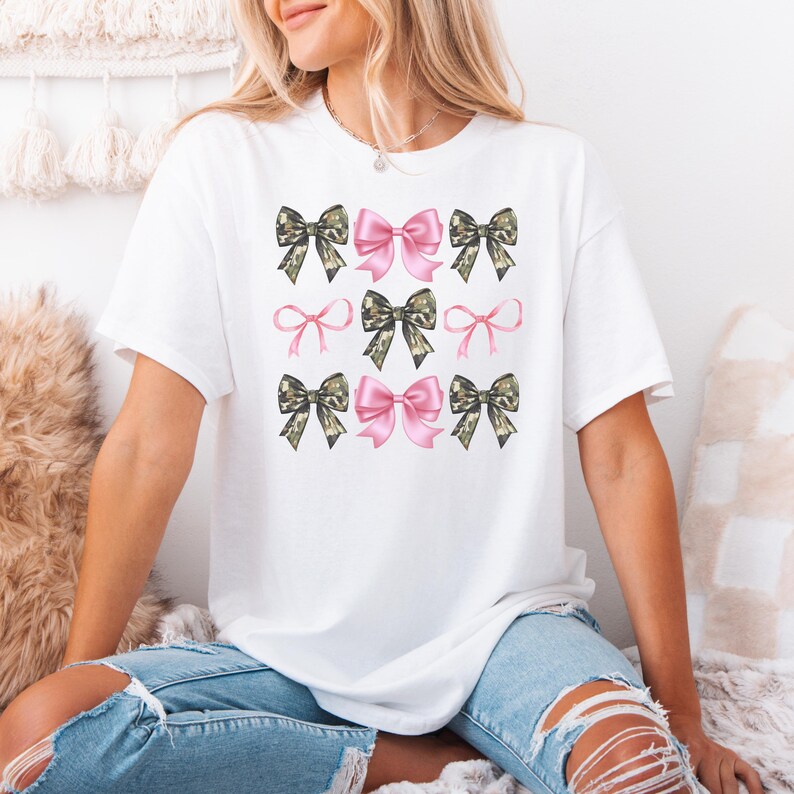 May include: A white short-sleeved t-shirt featuring a graphic print of nine bows arranged in a grid. The design alternates between solid pink bows and camouflage-patterned bows in shades of green, brown, and black, creating a repeating pattern.