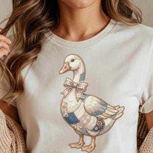 May include: A cream-colored t-shirt featuring a whimsical goose illustration. The goose has a patchwork design in shades of blue, pink, and floral patterns, with a plaid bow. The artwork is detailed and charming, perfect for casual wear.