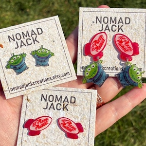 May include: Three sets of acrylic earrings on branded Nomad Jack display cards. The designs feature green three-eyed aliens and red rocket-shaped Pizza Planet logos. The earrings are presented as stud and dangle styles with clear, glossy finishes.