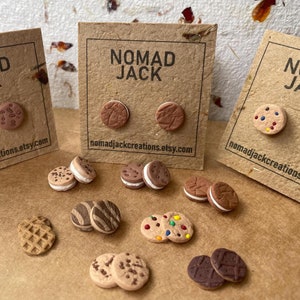 May include: A set of handmade polymer clay earrings featuring various cookie designs, including chocolate chip, sprinkle, and sandwich cookies. The earrings are displayed on brown card stock with the text "Nomad Jack" and a website address.