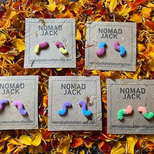 May include: Six pairs of colorful gummy worm earrings on brown cards with the text "NOMAD JACK" and the website nomadjackcreations.etsy.com. The earrings are in the colors pink, yellow, blue, green, and purple.