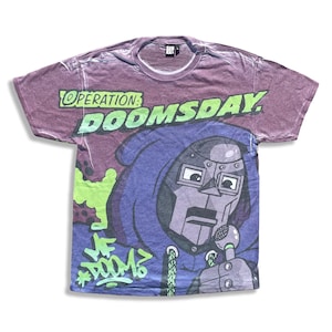 Puede incluir: Camiseta morada con un gráfico de un personaje enmascarado con un micrófono. La camiseta presenta el texto "OPERATION: DOOMSDAY" en verde y "MF DOOM" en estilo graffiti.