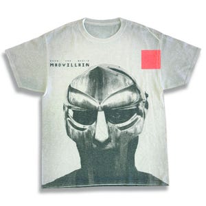 以下が含まれることがあります： マスクの白黒イラストが描かれたライトグレーのTシャツ。「DOOM AND MADVILLAIN」の文字がマスクの上にプリントされています。右上に小さな赤い正方形があります。