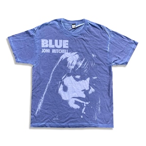 May include: Blue short-sleeve t-shirt featuring a portrait of Joni Mitchell. The shirt has the word "BLUE" printed above the singer's image, with "JONI MITCHELL" printed below. The shirt is a faded blue color.