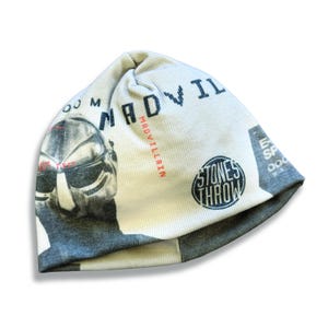 Puede incluir: Un gorro blanco y gris con un gráfico de una persona con gafas de sol y el texto "MADVILLAIN" y "STONES THROW". El gorro está hecho de un material suave y tiene un borde gris.