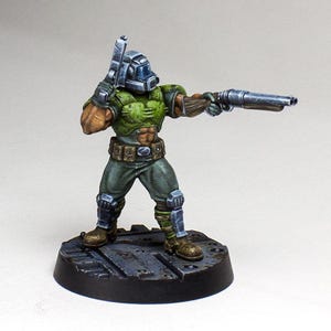May include: A green and gray painted miniature figure of a soldier holding a gun. The figure is standing on a round base with a textured surface.