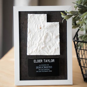May include: A framed white relief map of Utah with a small gold pin. Below the map is a black plaque with the name "Elder Taylor" and text in Spanish. The frame is white, set against a dark background.