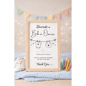May include: A framed sign with the text "Decorate a Bib or Onesie" in elegant script. The sign features illustrations of a baby onesie and bib hanging on a clothesline, with the words "Thank You" at the bottom. The frame is light wood.