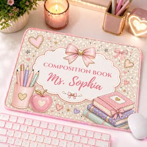 Pu&ograve; includere: Un tappetino per mouse rosa e beige personalizzato con il testo "COMPOSITION BOOK Ms. Sophia". Il design include cuori, fiocchi, matite e libri. Una tastiera e un mouse rosa sono in primo piano.