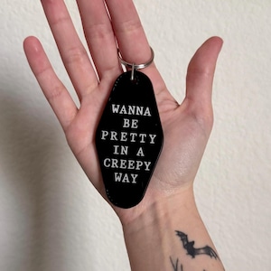 May include: Black keychain with the text "WANNA BE PRETTY IN A CREEPY WAY" in white. The keychain is a retro motel style with a silver key ring.
