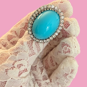 May include: An oval-shaped brooch featuring a large, smooth, bright turquoise cabochon stone at the center. The turquoise is surrounded by a border of small, clear, sparkling round-cut stones set in a metallic frame with a polished silver finish.