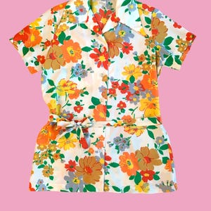 May include: A vintage floral print romper set. The set includes a short-sleeved button-down shirt and matching shorts, both featuring a vibrant pattern of orange, yellow, red, and brown flowers on a white background. A fabric belt cinches the waist.