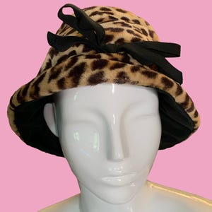 May include: A leopard print cloche hat with a black ribbon bow. The hat features a faux fur texture in shades of tan and brown, with a black interior lining. The hat is displayed on a white mannequin head against a pink background.