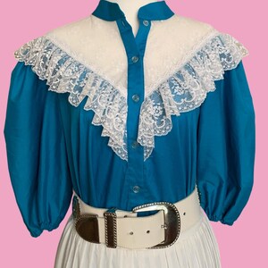 May include: A vintage teal blouse with a white lace yoke and ruffled trim. The blouse has short, puffed sleeves and a button-up front. A white belt with a silver buckle cinches the waist, paired with a white pleated skirt. The outfit is displayed on a mannequin.