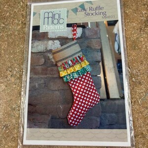 May include: A sewing pattern for an Easy Ruffle Stocking by Mini Mac Patterns. The image shows a red stocking with white polka dots, a burlap cuff, and colorful ruffles. The pattern is displayed in a clear plastic sleeve.