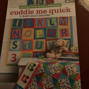 May include: A book titled "Cuddle Me Quick" with 11 baby quilt designs. The cover features a colorful alphabet quilt with a teddy bear and a toy car.