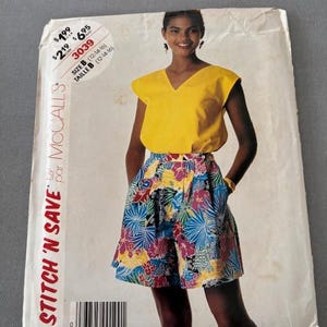 May include: A vintage McCall's sewing pattern featuring a woman wearing a yellow top and floral shorts. The pattern is labeled "STITCH 'N SAVE" and includes size and price information. The shorts have a vibrant pattern with blue, pink, and yellow flowers.