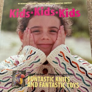 Puede incluir: Portada de libro con el título "Kids Kids Kids" en rosa. La portada presenta a un niño sonriente que lleva un suéter color crema con detalles de punto coloridos. El libro trata sobre patrones de tejido y juguetes divertidos.