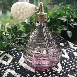 Puede incluir: Un frasco de perfume vintage de vidrio con un atomizador de bulbo blanco. El frasco es de color rosa claro y tiene un diseño texturizado en forma de cuadrícula.