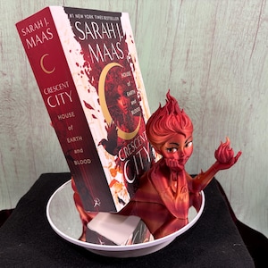 May include: A red and orange figurine of a woman with fiery hair and a flame in her hand, holding a book titled "Crescent City: House of Earth and Blood" by Sarah J. Maas. The book is propped up in a white bowl.